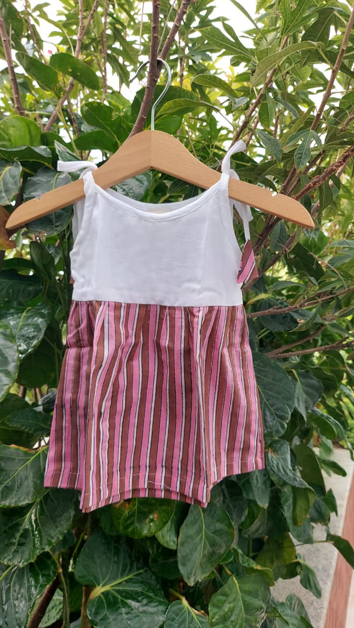 A toddler girl's sleeveless romper hanging on a wooden hanger against a background of green garden foliage. The romper features a solid white top half and a pink and white vertically striped bottom half.