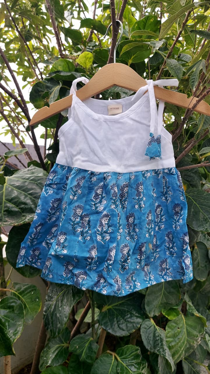 A toddler girl's sleeveless top and skirt set hanging on a wooden hanger outdoors against a background of green leaves. The top is white with thin shoulder straps, and the skirt is blue with a white floral pattern.