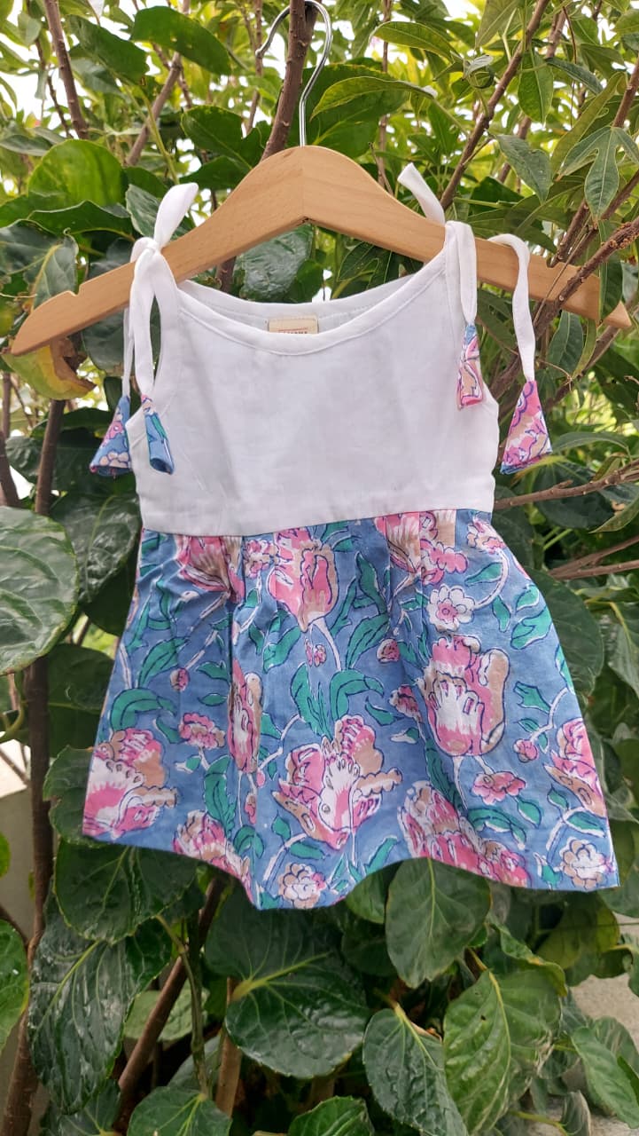 A toddler girl's knot dress hanging on a wooden hanger against a background of green leaves. The top features a solid white upper bodice and a blue floral skirt with pink and green tulip patterns.