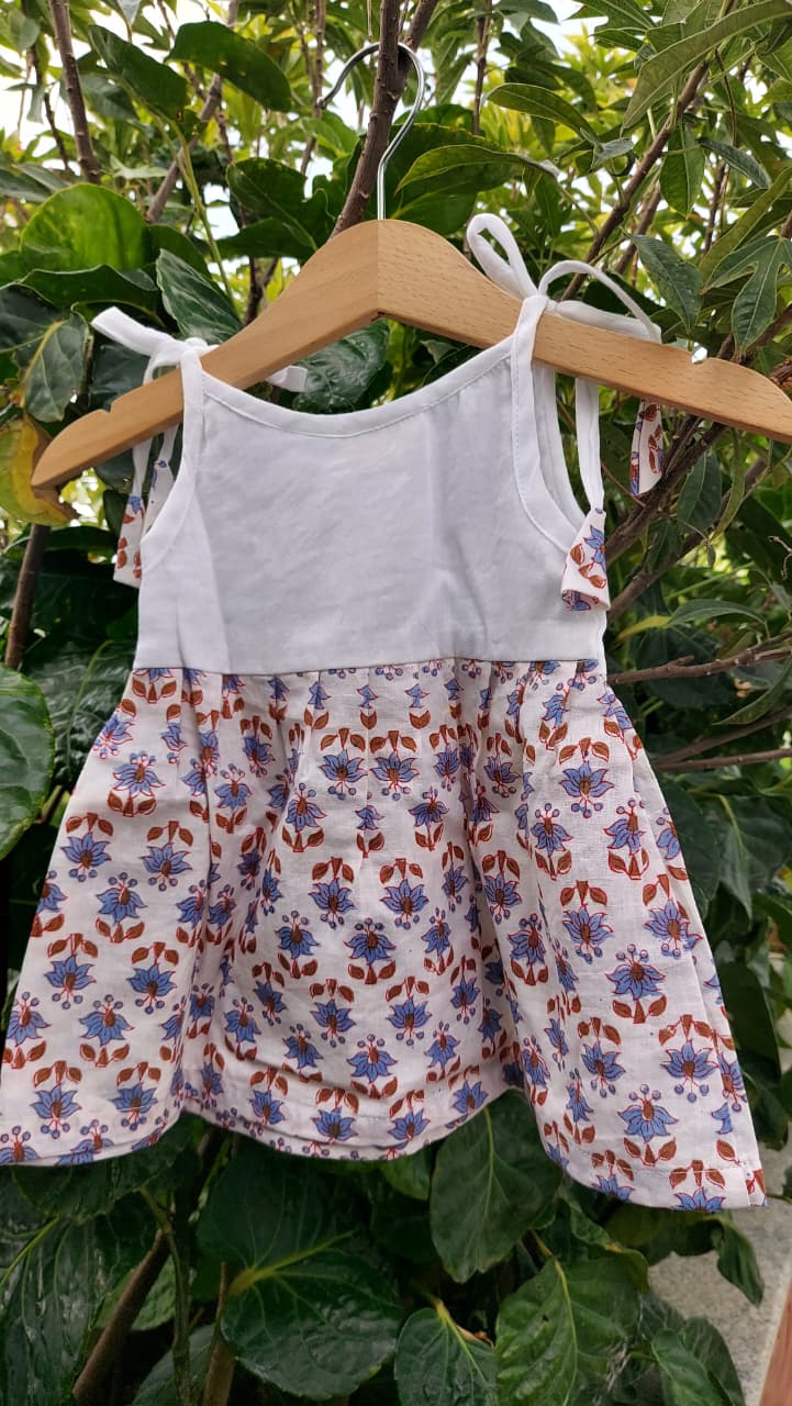 A toddler girl's sleeveless dress hanging on a wooden hanger against a background of green garden foliage. The dress features a solid white upper bodice and a white skirt decorated with a colorful floral pattern in purple, blue, and orange.
