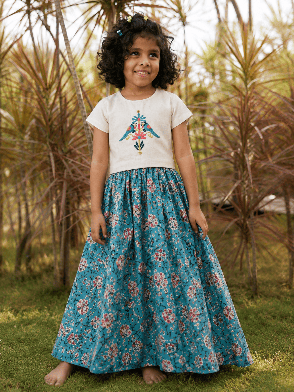 A young girl with curly dark hair stands barefoot outdoors in a grassy area with palm trees, wearing a white short-sleeve top with a colorful embroidered bird design and a long teal floral skirt.