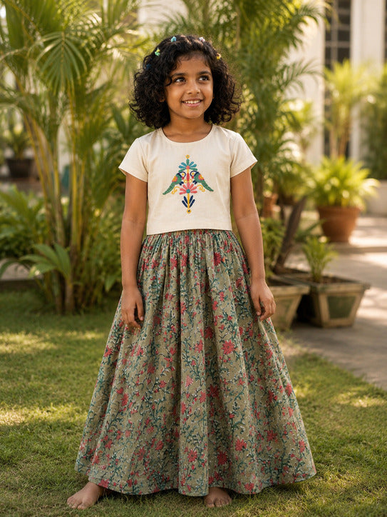 A young girl with curly black hair stands barefoot on a grassy lawn wearing a two-piece outfit. The outfit consists of a cream-colored short-sleeve top with a colorful embroidered bird design on the chest, and a long, flared skirt in a muted sage green with a red and pink floral pattern.