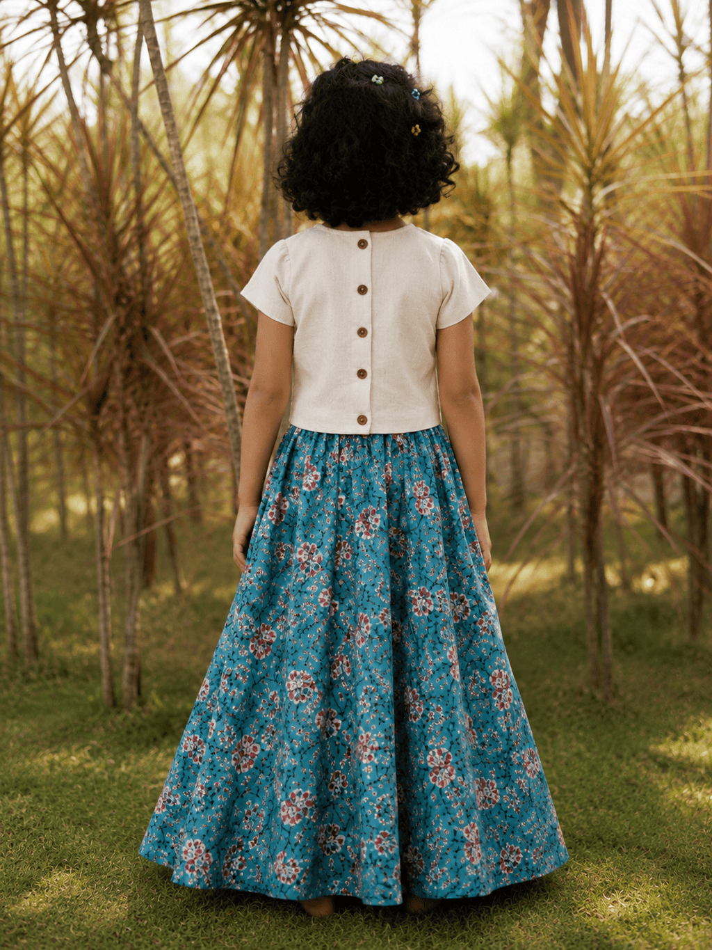 A child viewed from behind wearing a cream short-sleeve top with brown buttons and a teal blue floral skirt, standing in a grassy outdoor area with palm trees.