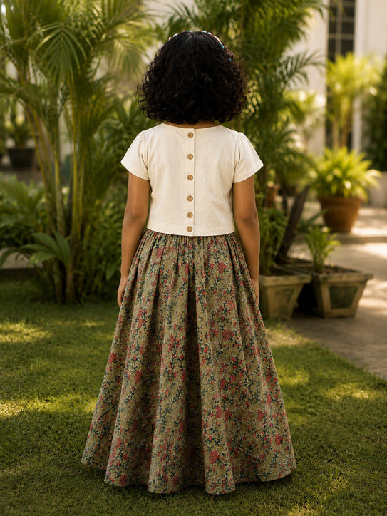 A child viewed from behind wearing a cream short-sleeve top with a button back and a long floral skirt, standing on grass in a garden with potted plants.
