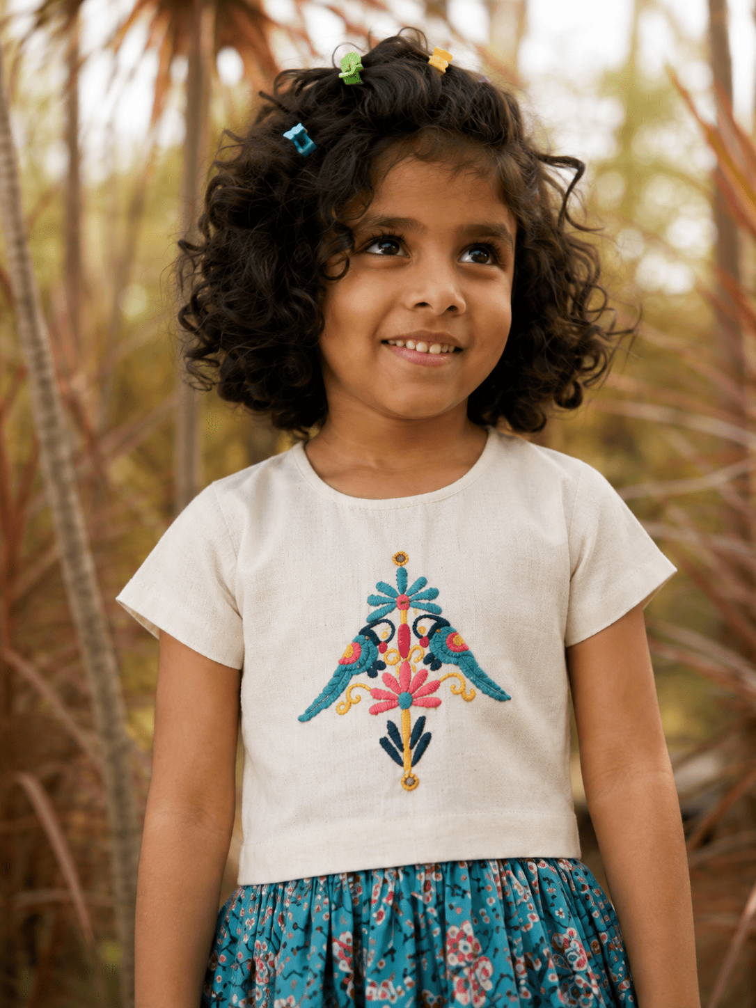 A young girl with curly dark hair wearing a cream-colored short-sleeve top with colorful embroidered birds and a blue floral skirt, standing outdoors against a blurred background of trees.