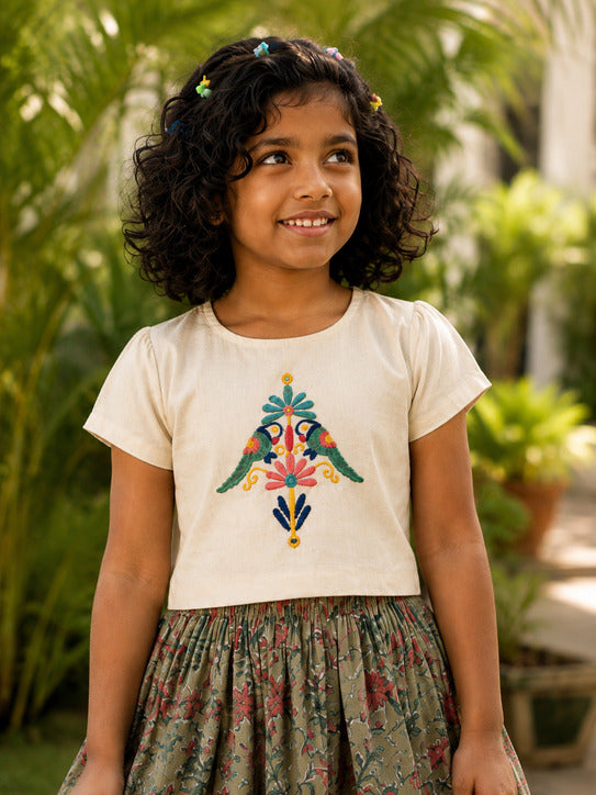 A young girl with curly dark hair is wearing a cream-colored short-sleeve top with a colorful embroidered bird motif on the chest, paired with a patterned green skirt, set against a lush green garden background.