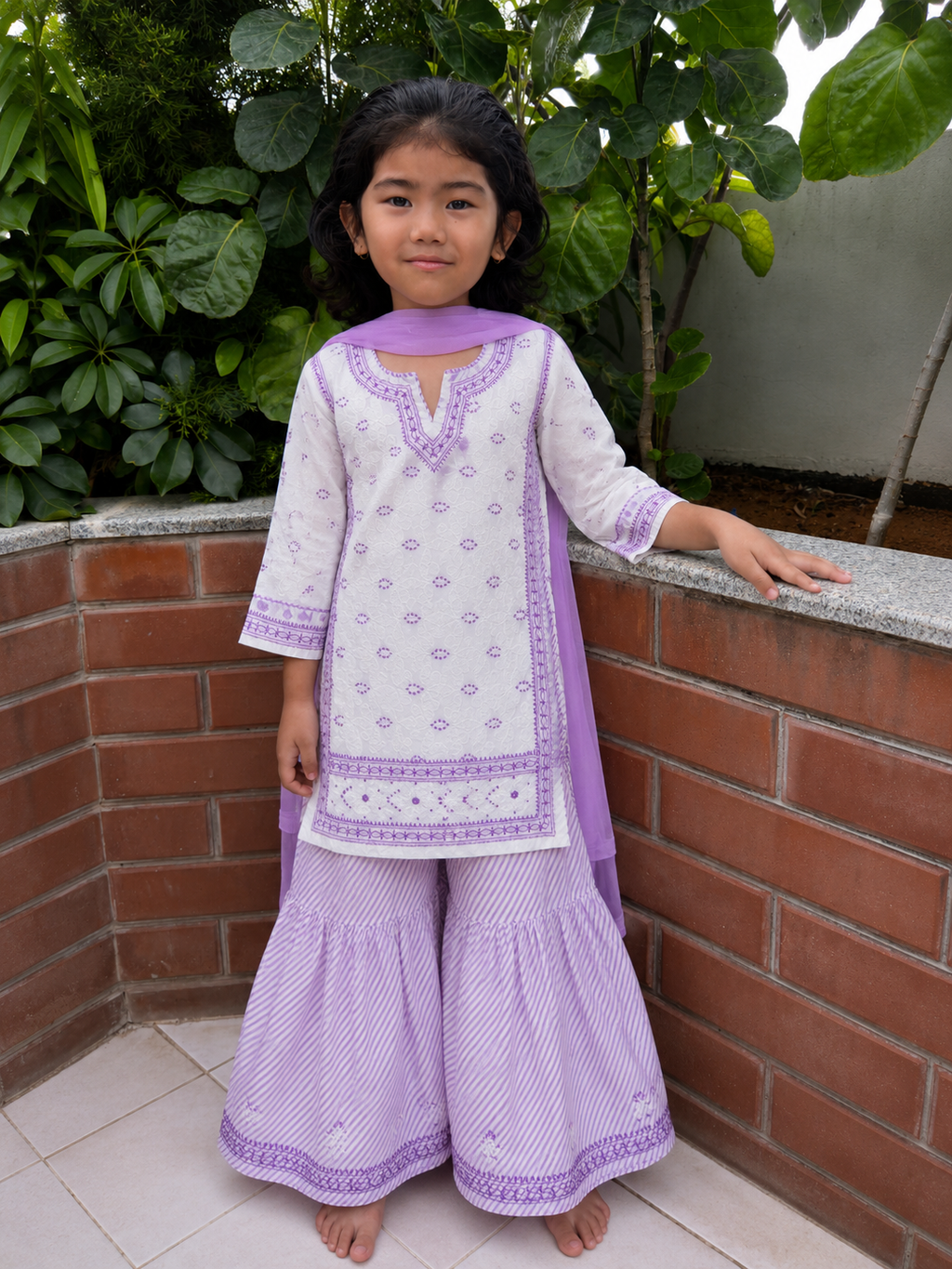 A young girl stands barefoot outdoors against a brick wall and green plants, wearing a white and light purple traditional outfit with matching patterned pants and a draped scarf.