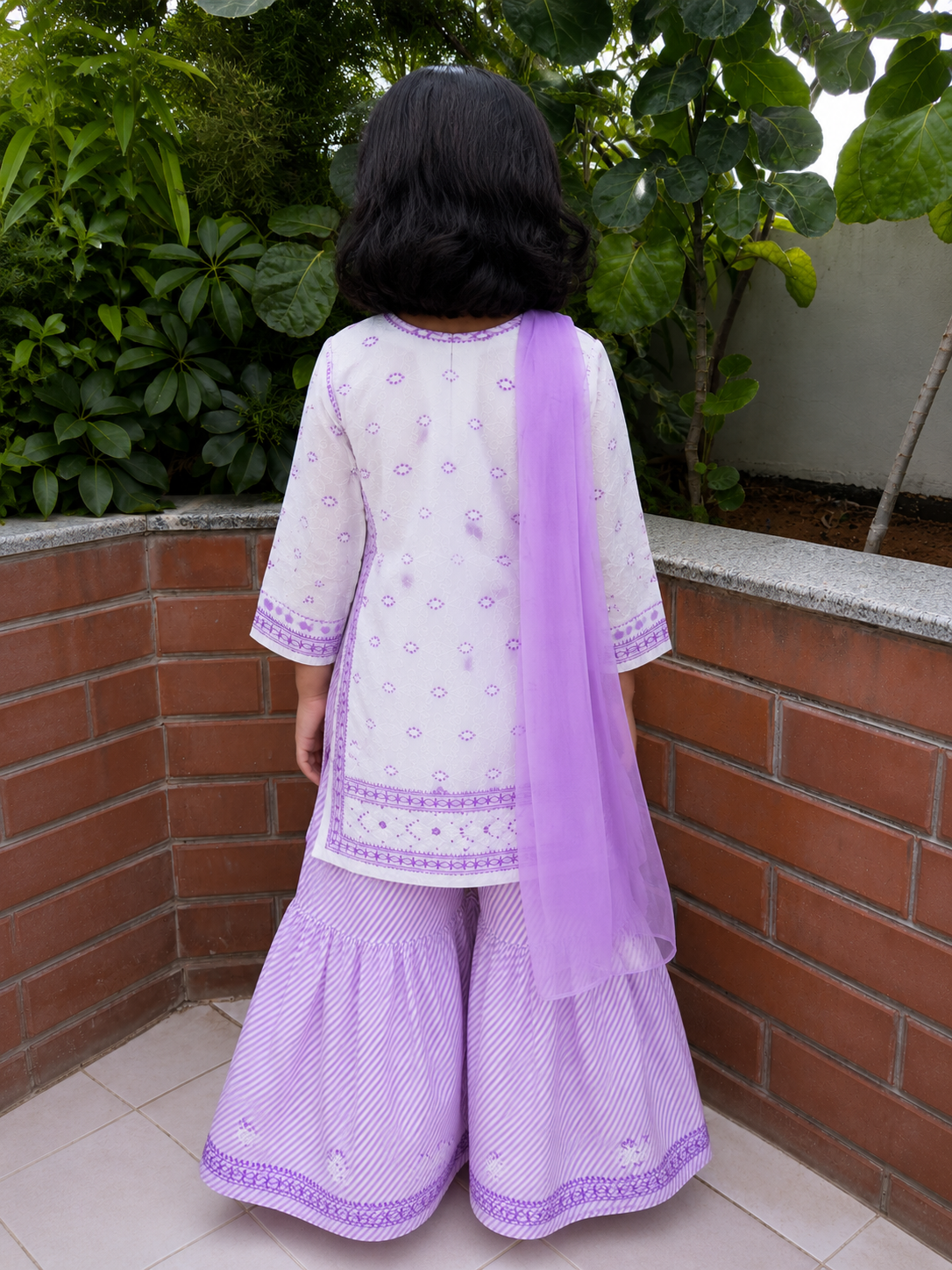 A child viewed from behind wearing a light purple and white traditional outfit with wide flared pants and a sheer purple dupatta, standing outdoors against a brick wall and green plants.