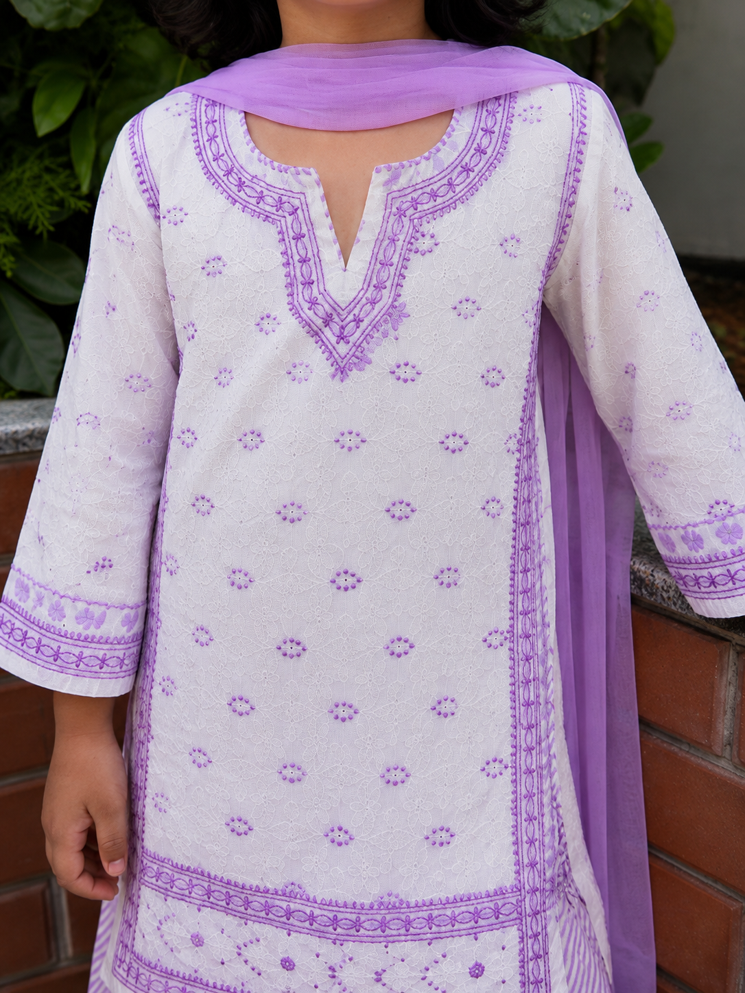A close-up of a child wearing a white kurta with purple Chikankari and a matching purple dupatta, shown outdoors against a brick wall and green plants.