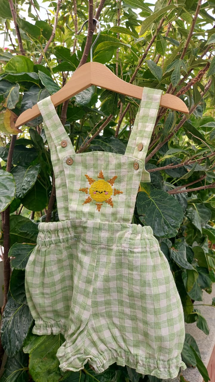 A baby romper in a light green and white gingham pattern, featuring an cross stitch embroidered yellow sun on the chest and adjustable shoulder straps, hanging on a wooden hanger against a background of green leaves.