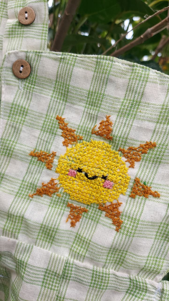 Close-up of a green and white checkered fabric garment, likely a bib or pocket cardigan, featuring a yellow embroidered sun with orange rays and a smiling face on the front.