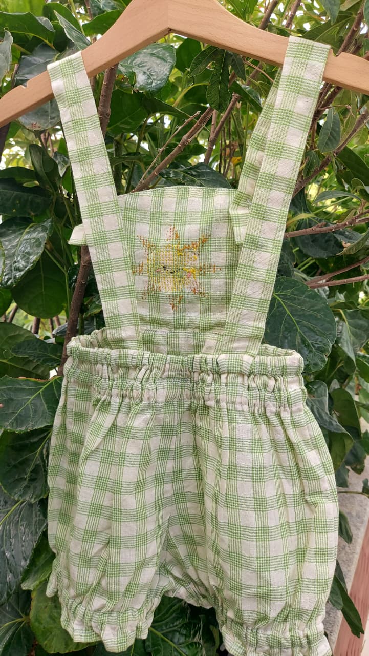 A baby romper in a green and white gingham check pattern, featuring an embroidered yellow sun on the chest, hanging on a wooden hanger against a background of green leaves.