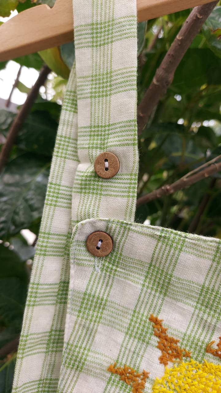 Close-up of a fabric tote bag strap with a green and white plaid pattern, two wooden buttons, and a partial embroidered sunflower motif, hanging against a blurred outdoor background of green leaves and branches.