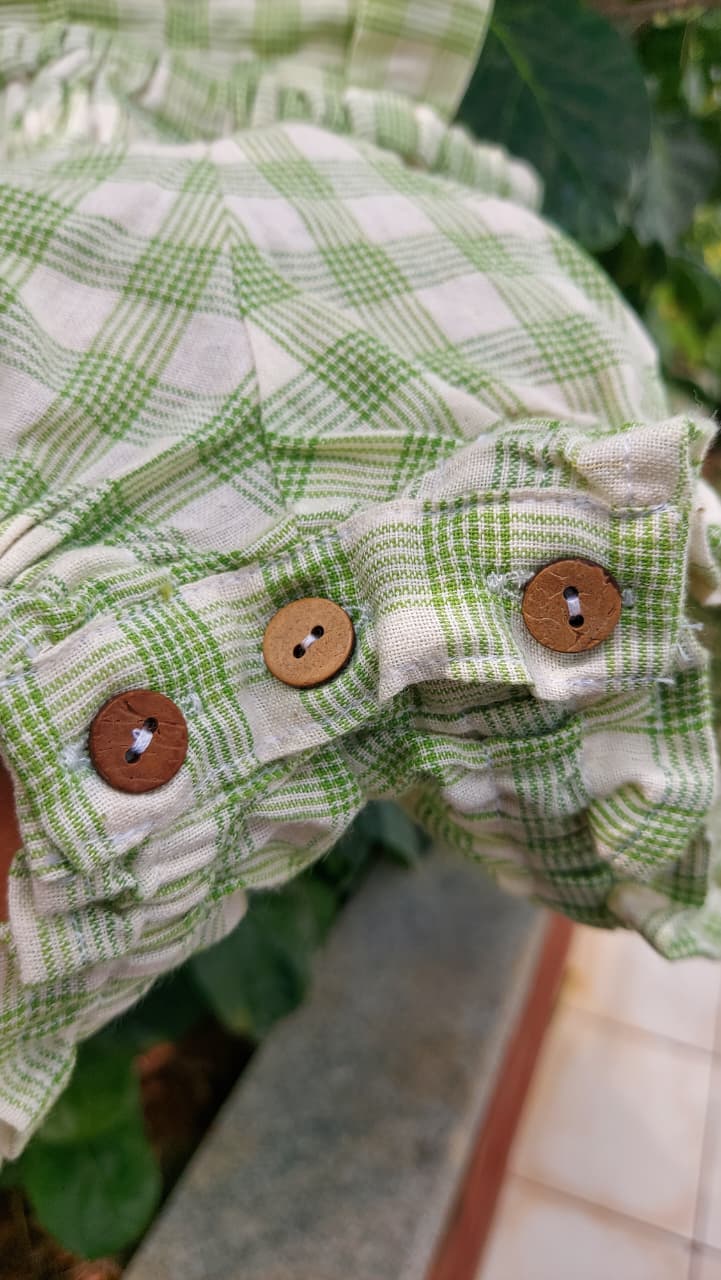 Close-up of a green and white gingham plaid garment with ruffled edges and three wooden buttons, photographed outdoors with foliage and a stone surface visible.