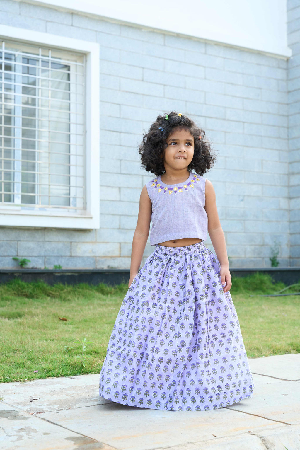 Young girl in a light purple top and floral skirt standing outdoors on a stone path.