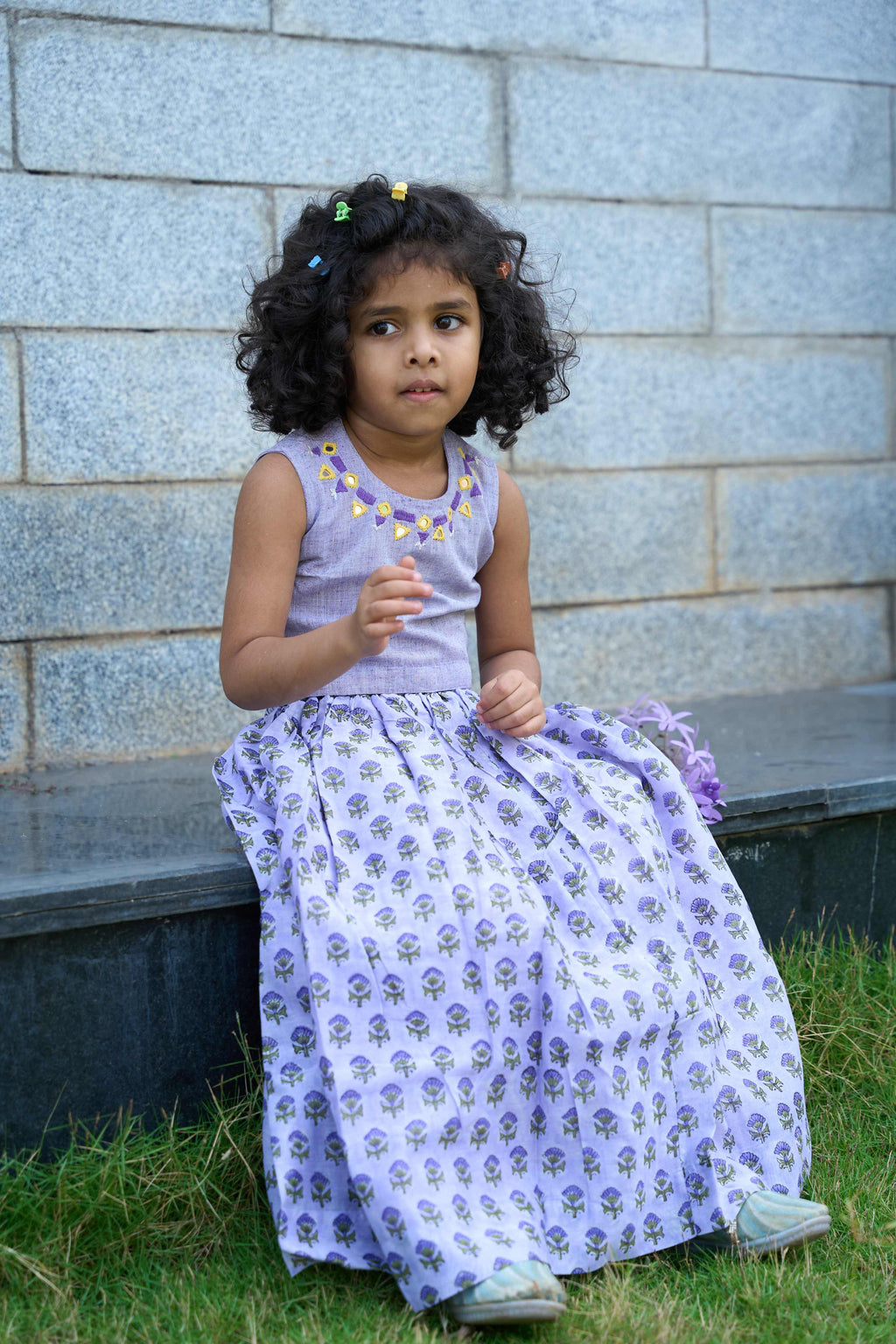 Young girl in a purple dress sitting on a bench against a stone wall.