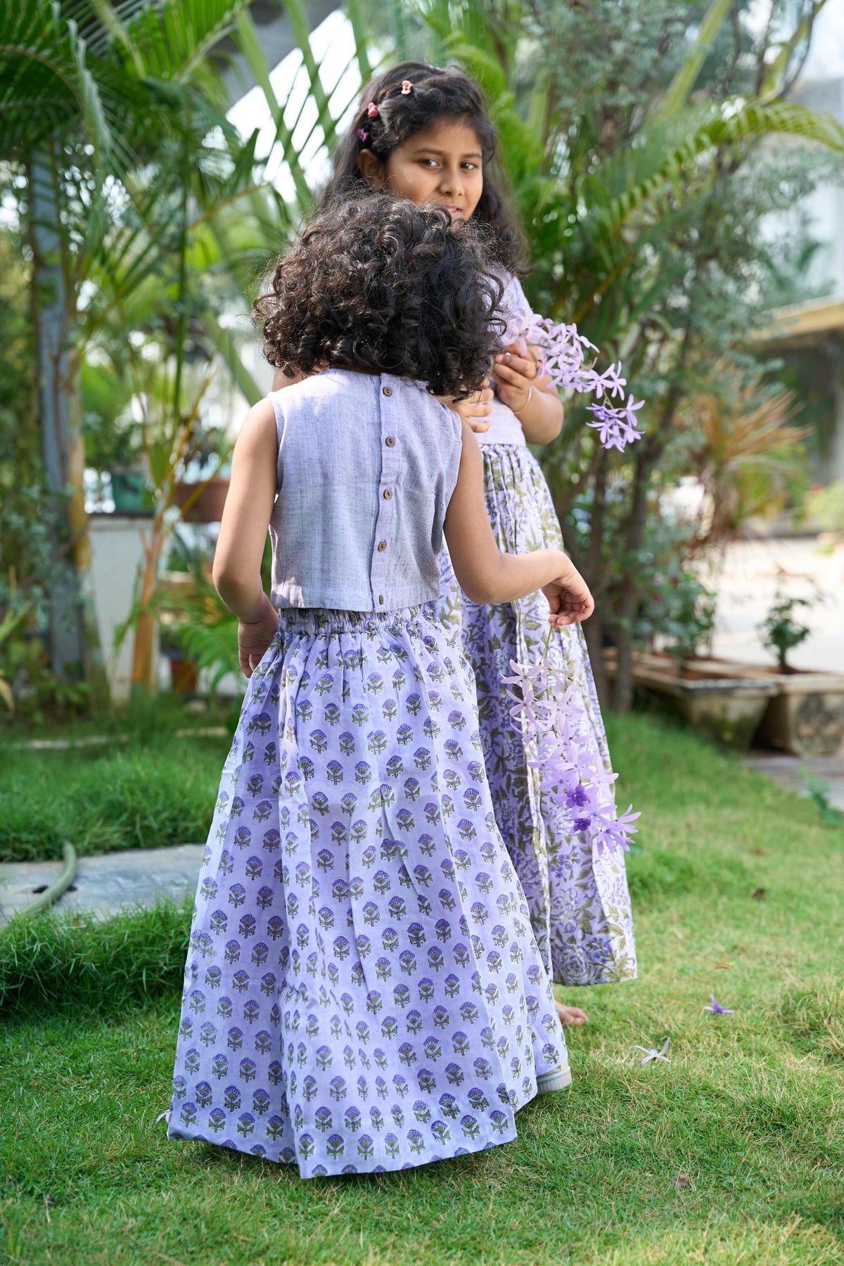 Two children in matching dresses standing in a garden with greenery around.