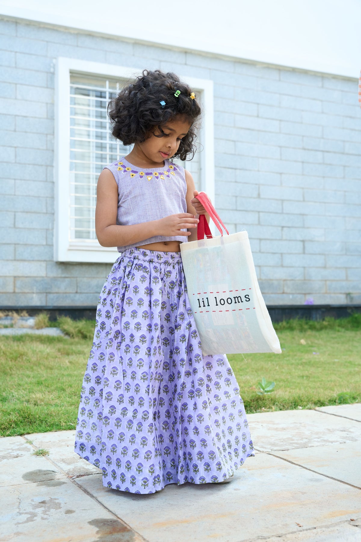 Young girl in a purple dress holding a 'lil looms' tote bag outdoors.