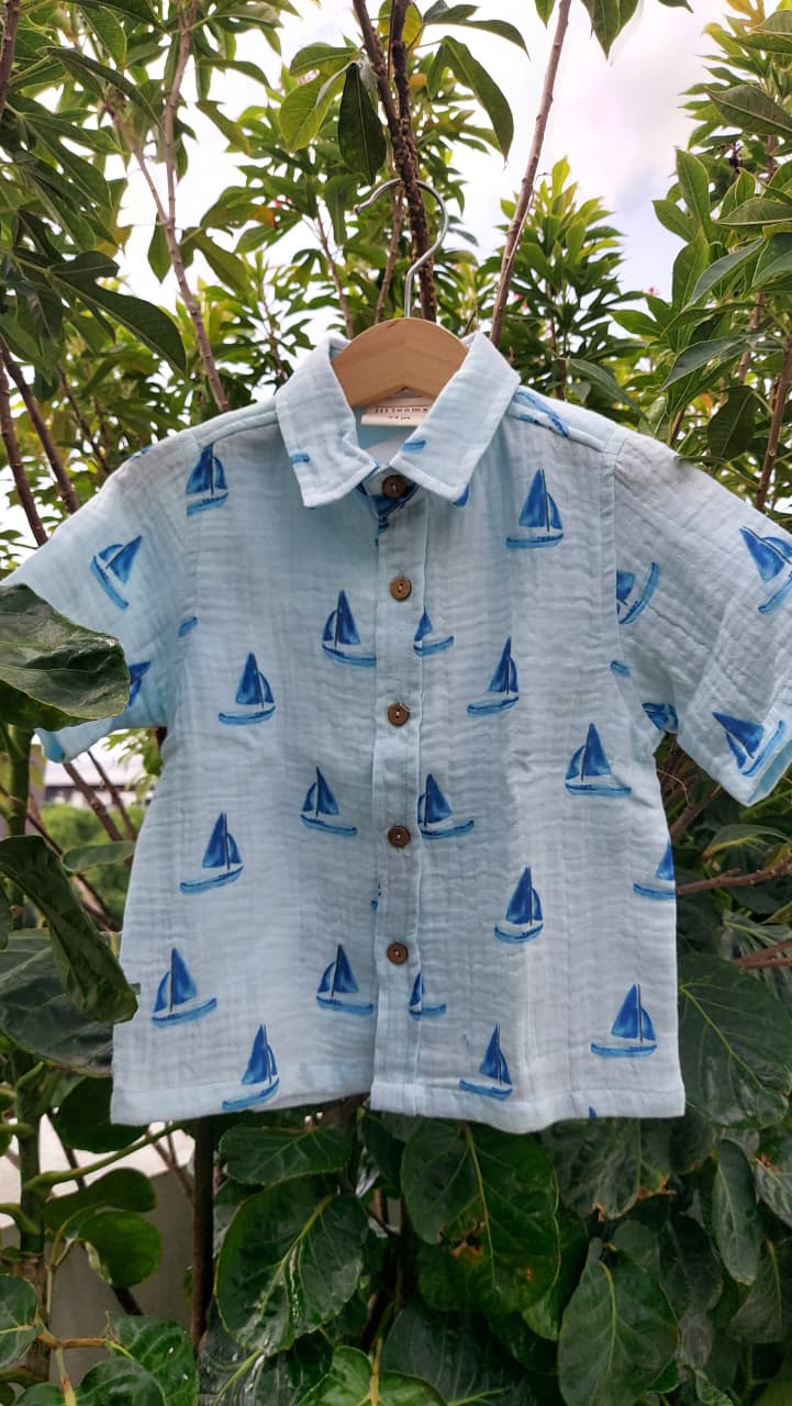 A light blue short-sleeve button-up shirt with a blue sailboat print, hanging on a wooden hanger against a background of green outdoor foliage.