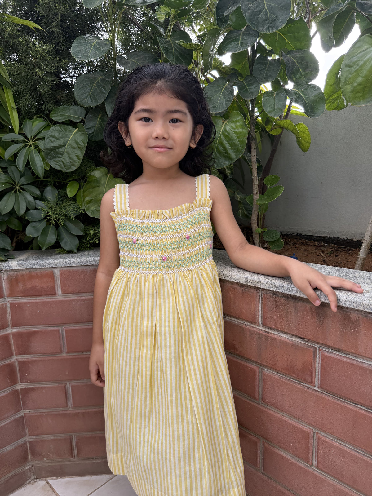 Young girl in a yellow dress standing outdoors near a brick wall and plants.
