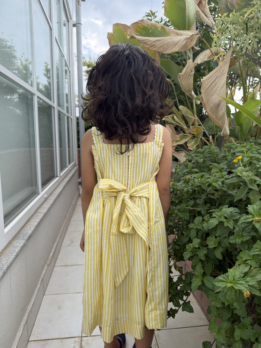 Child in a yellow striped dress standing near a building with plants.