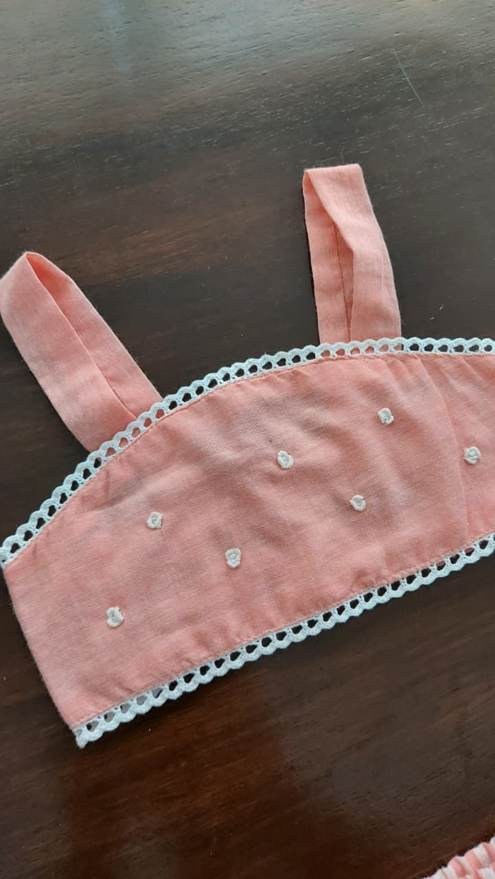 A pink fabric bikini top or bandeau-style top with white lace trim and small white embroidered dots, laid flat on a dark wooden surface.