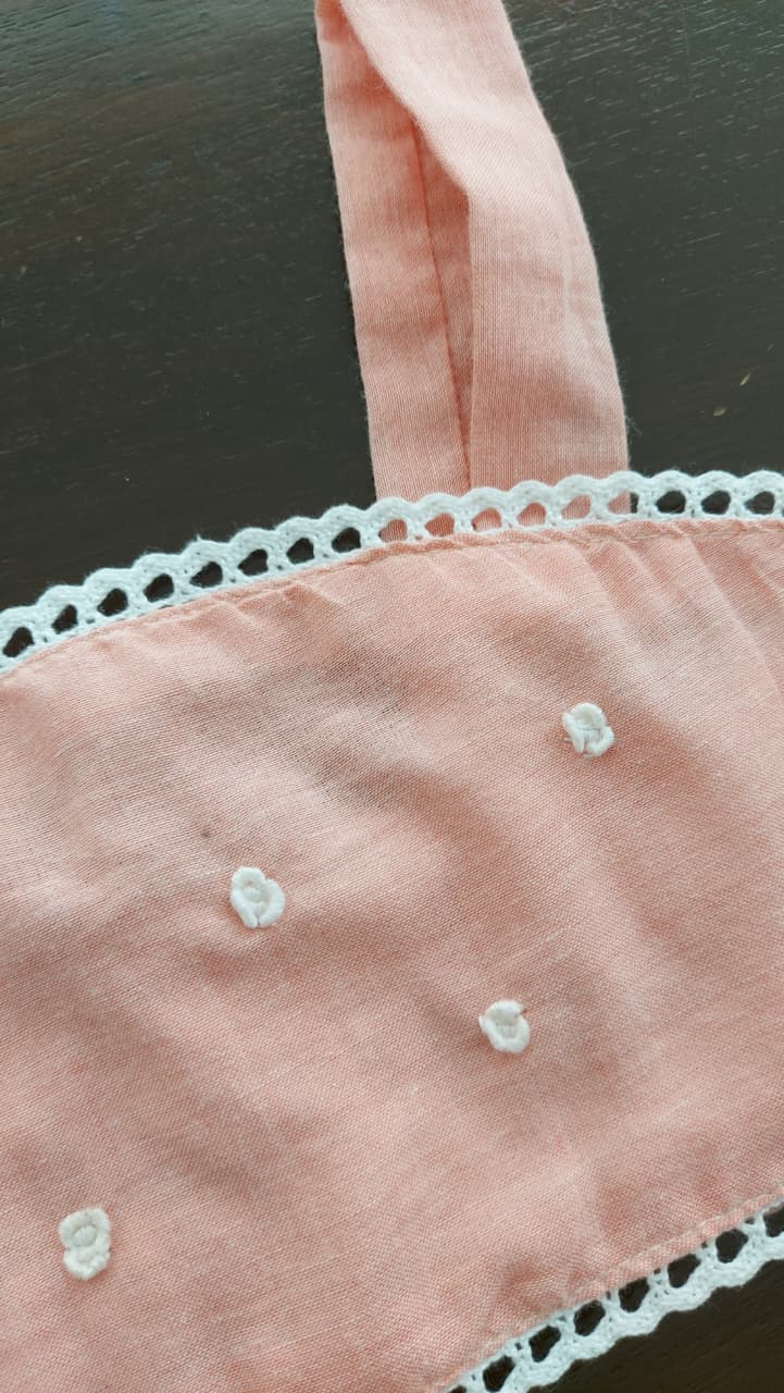 A close-up of a light pink Cotton soft fabric item, likely a bikini top or dress panel, featuring white lace trim along the neckline and small white embroidered flower details.