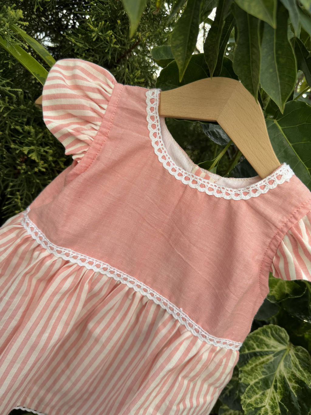 Pink and white striped dress with lace trim on a wooden hanger against a green leafy background