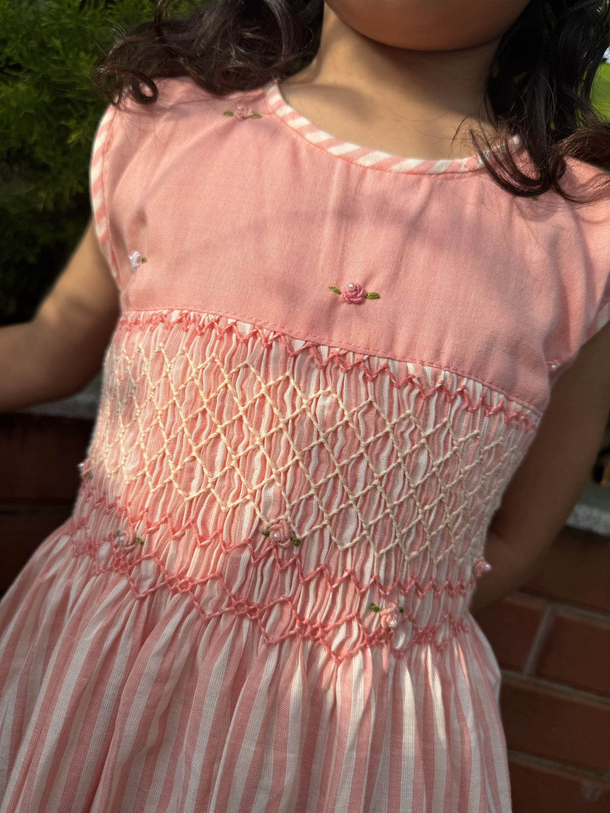A close-up of a child wearing a pink smocked dress outdoors. The dress has a solid pink bodice with small embroidered floral details, a smocked mesh waist panel, and a pink-and-white striped skirt.