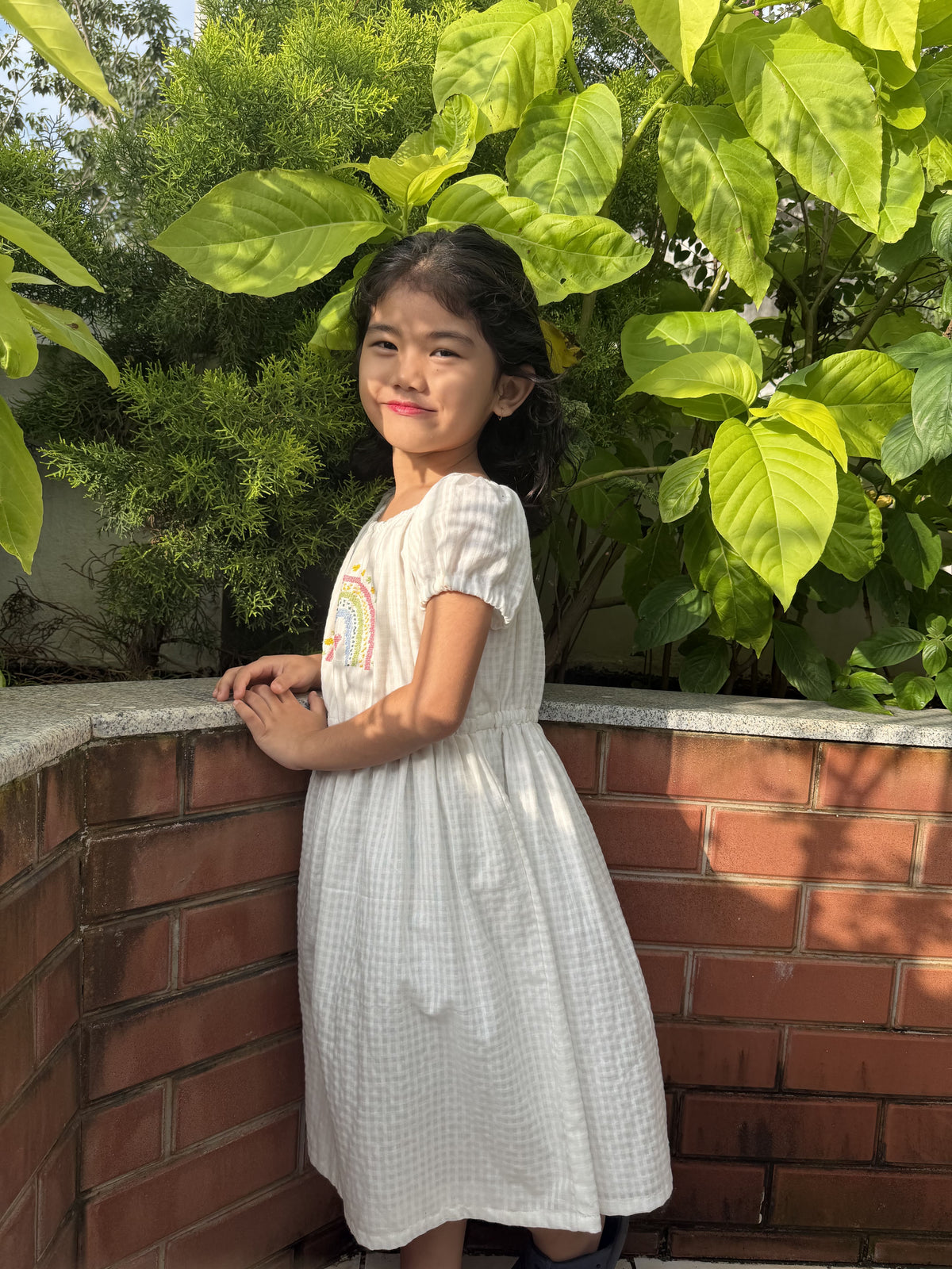 A young girl with dark hair stands outdoors against a brick wall and green foliage, wearing a white textured short-sleeve dress with a small rainbow graphic on the chest.