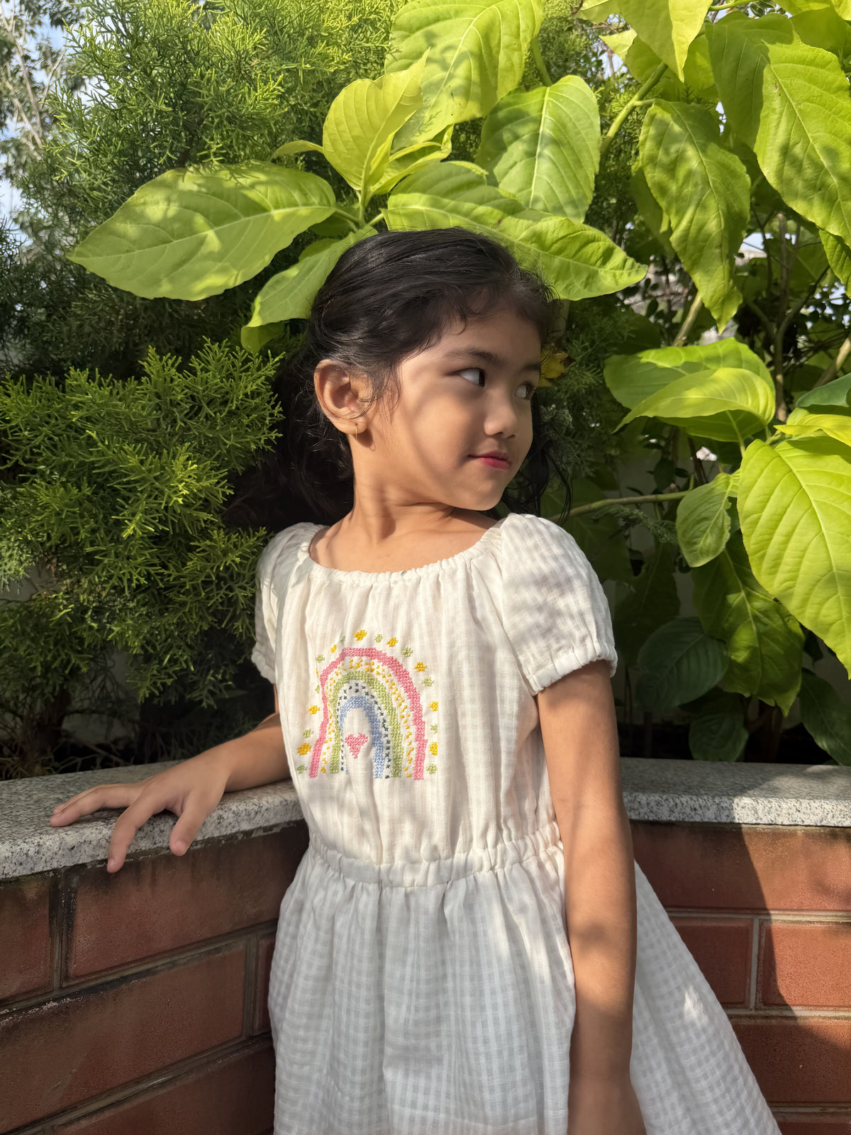 A young girl wearing a white and gray gingham dress with a colorful rainbow embroidery on the chest, standing outdoors next to a brick wall with green foliage.
