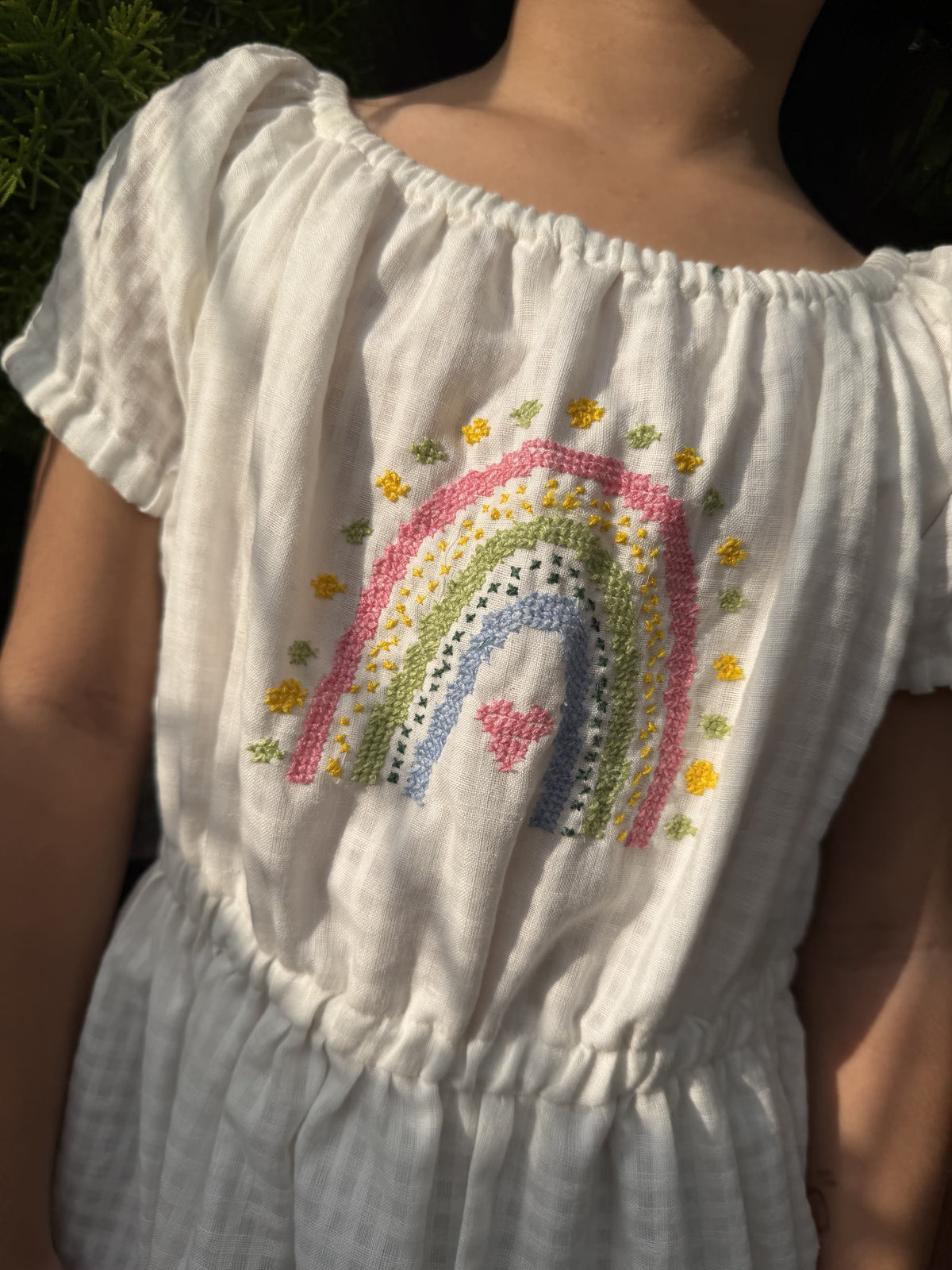 A close-up of a child wearing a white linen dress with a colorful embroidered rainbow and small yellow flowers on the chest.