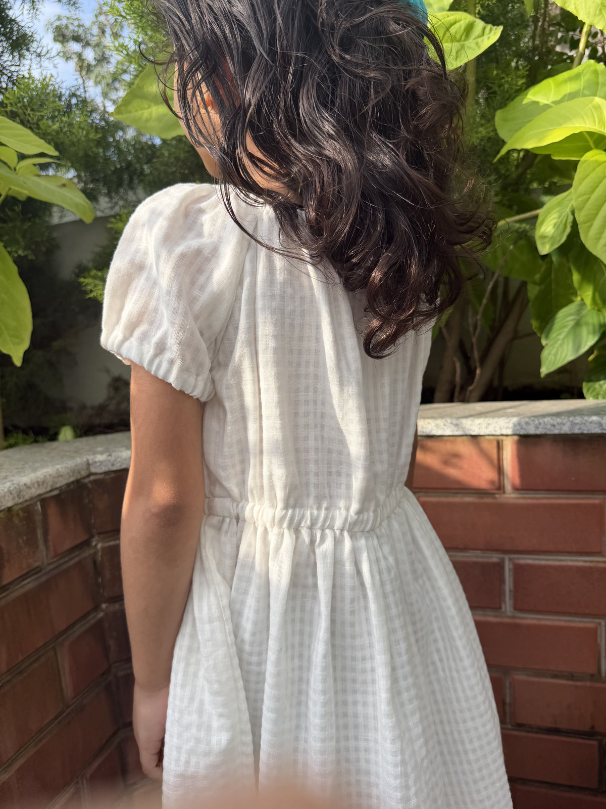 A child wearing a white short-sleeve dress with a subtle grid pattern, photographed from behind in an outdoor setting with brick walls and green foliage.