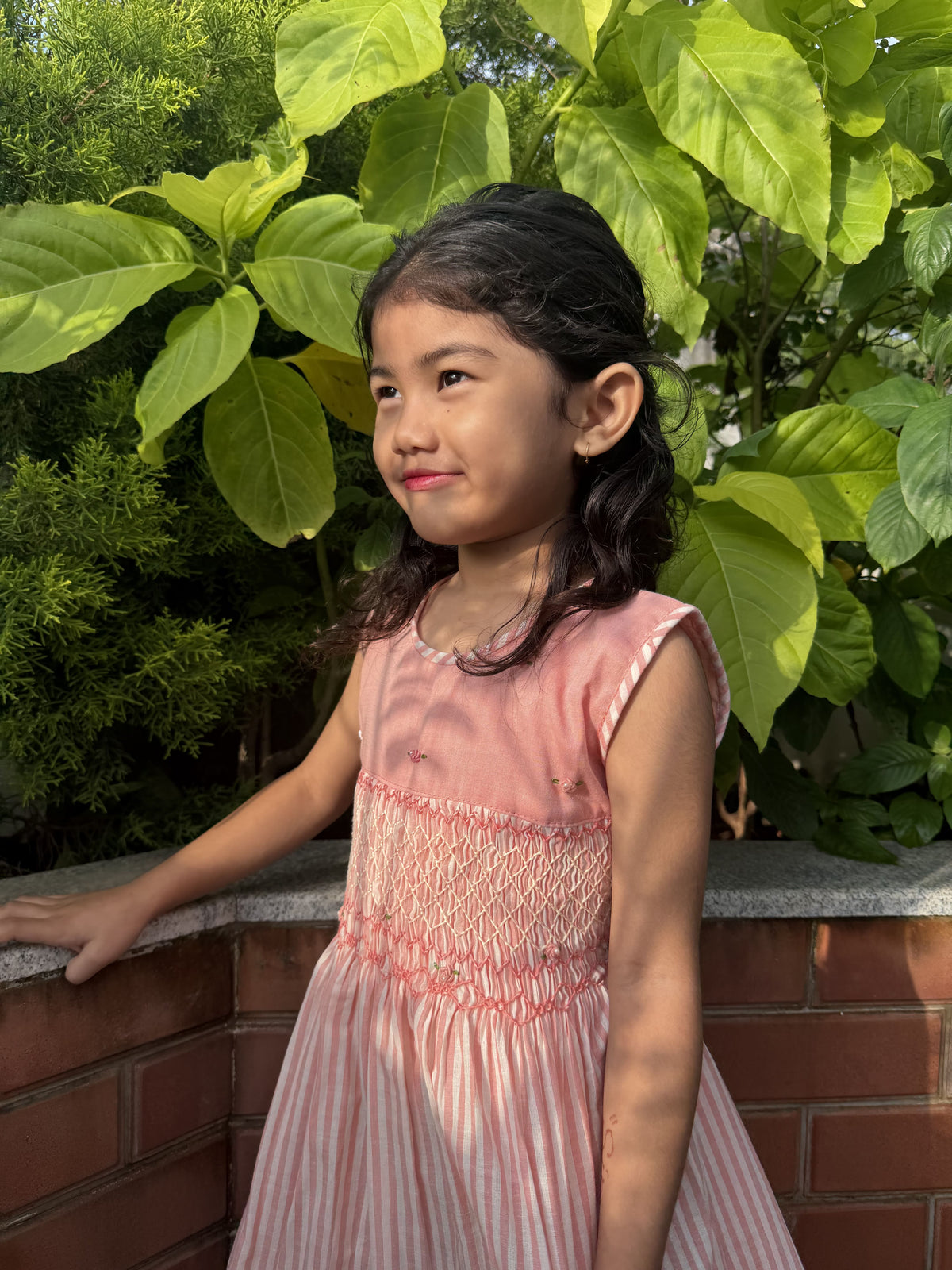 A young girl with dark hair wearing a pink smocked dress, standing outdoors next to a brick wall with green foliage in the background.