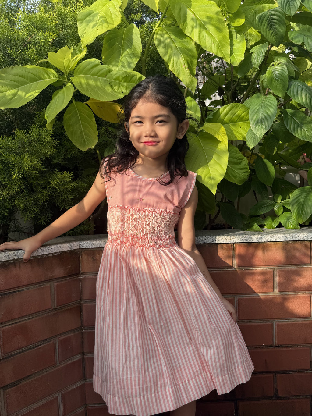 A young girl with dark hair stands outdoors against a brick wall and green foliage, wearing a sleeveless pink dress with vertical white stripes and smocked detailing at the bodice.