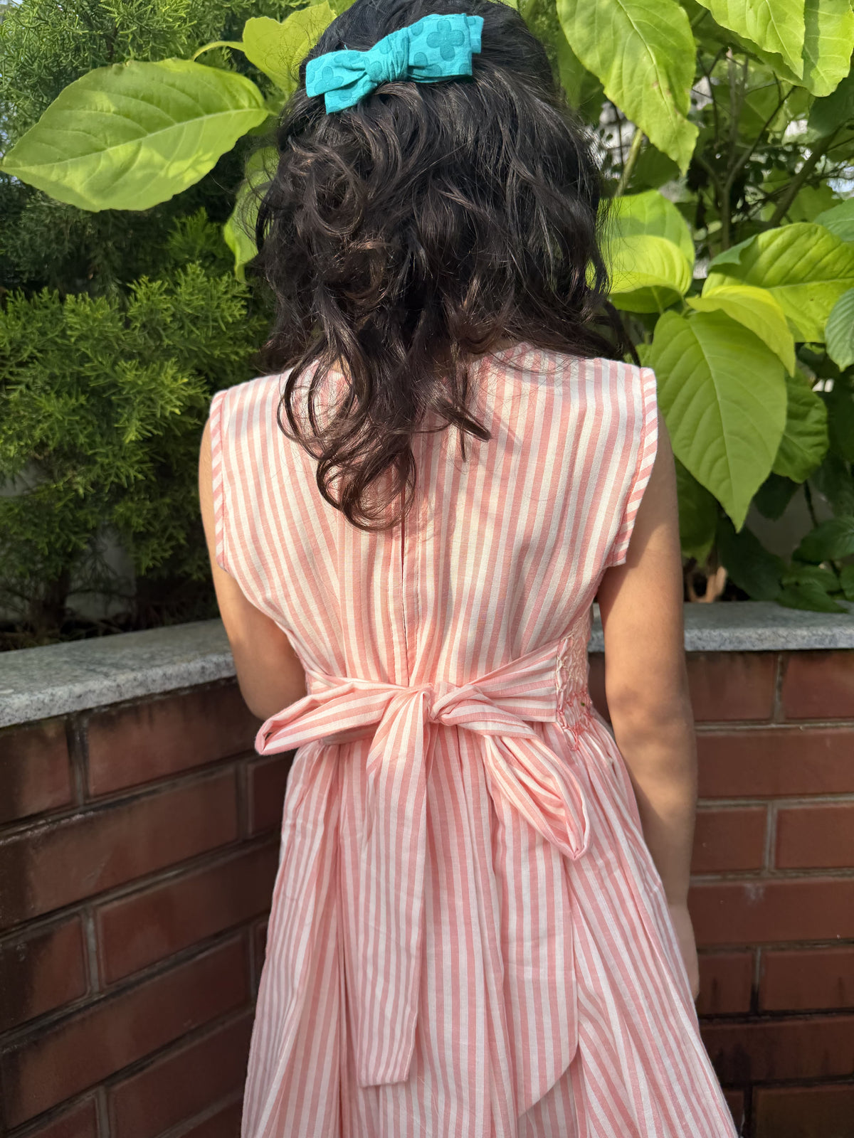 A child viewed from behind wearing a sleeveless pink and white vertically striped dress with a tied waist bow. The child has dark curly hair and wears a turquoise bow headband, standing outdoors against a brick wall and green foliage.