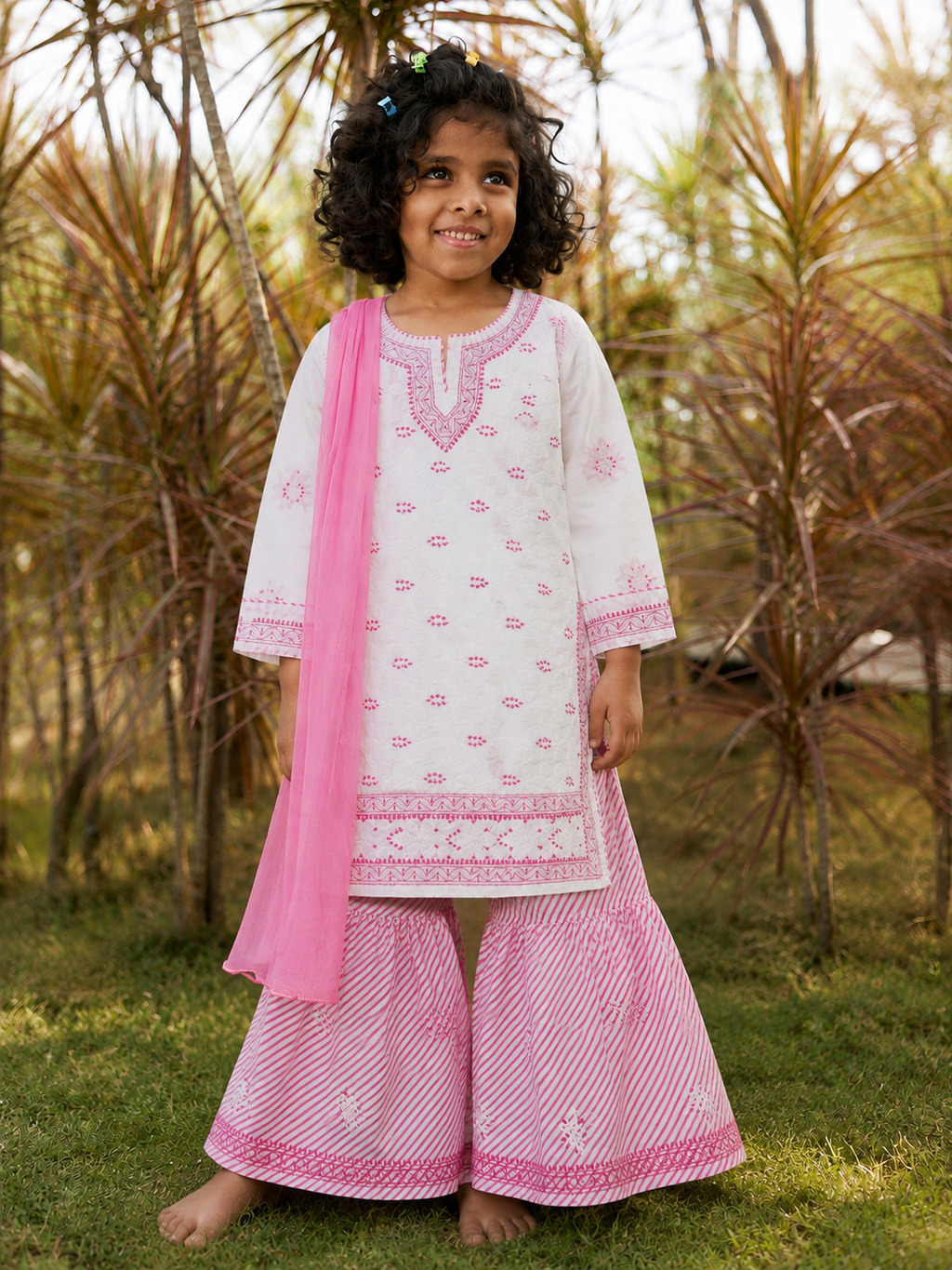 A young girl stands barefoot outdoors on grass in front of palm trees, wearing a white and pink traditional outfit with embroidered details, wide-leg pink patterned pants, and a pink dupatta.