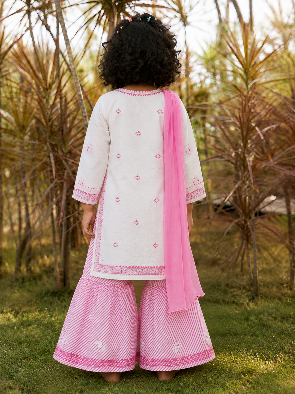 A child viewed from behind wearing a traditional outfit consisting of a white kurta with pink embroidery, wide pink and white striped flared pants, and a pink dupatta, standing outdoors in a garden.