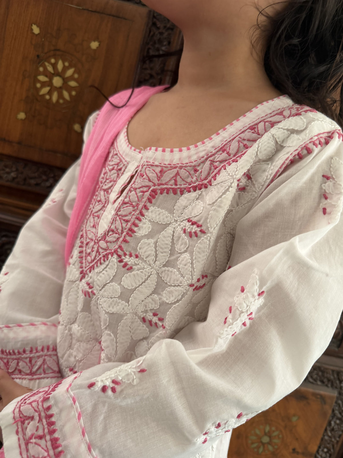 A close-up of a person wearing a white embroidered kurta with pink floral embroidery and a pink dupatta, set against a warm wooden background.