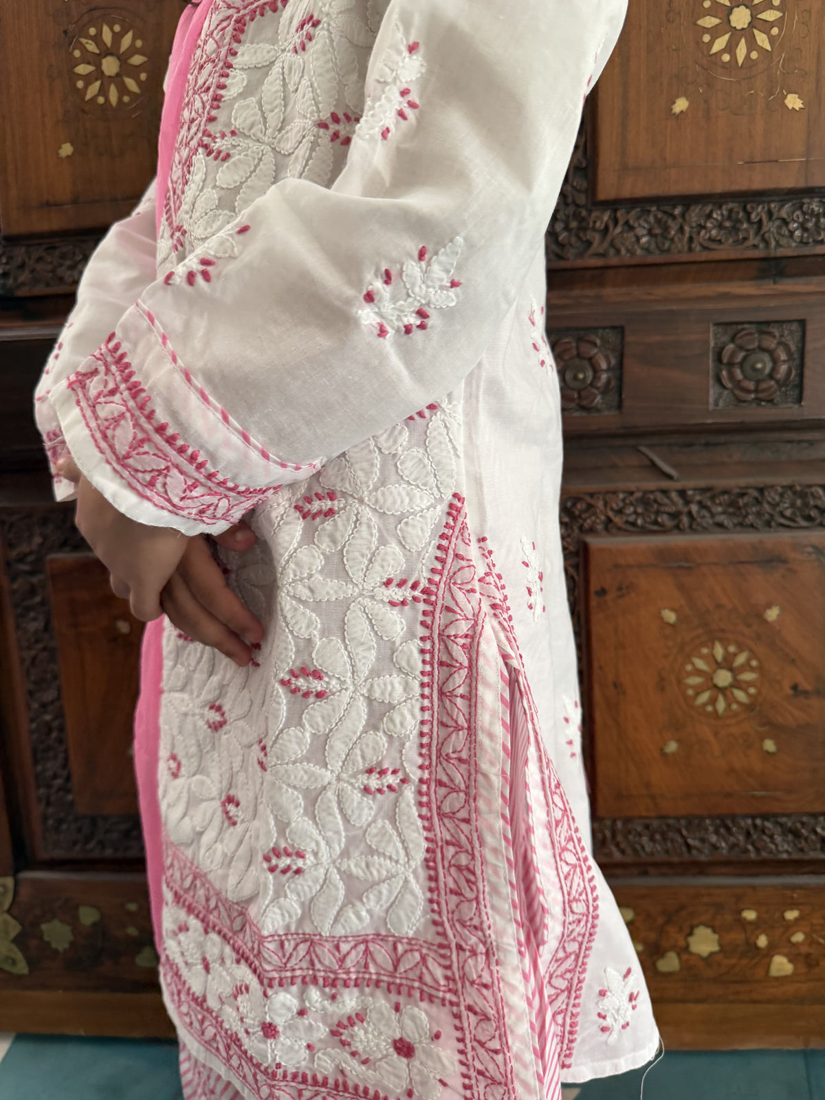 A close-up of a white embroidered kurta with pink border and floral embroidery, worn by a model standing in front of a carved wooden cabinet.