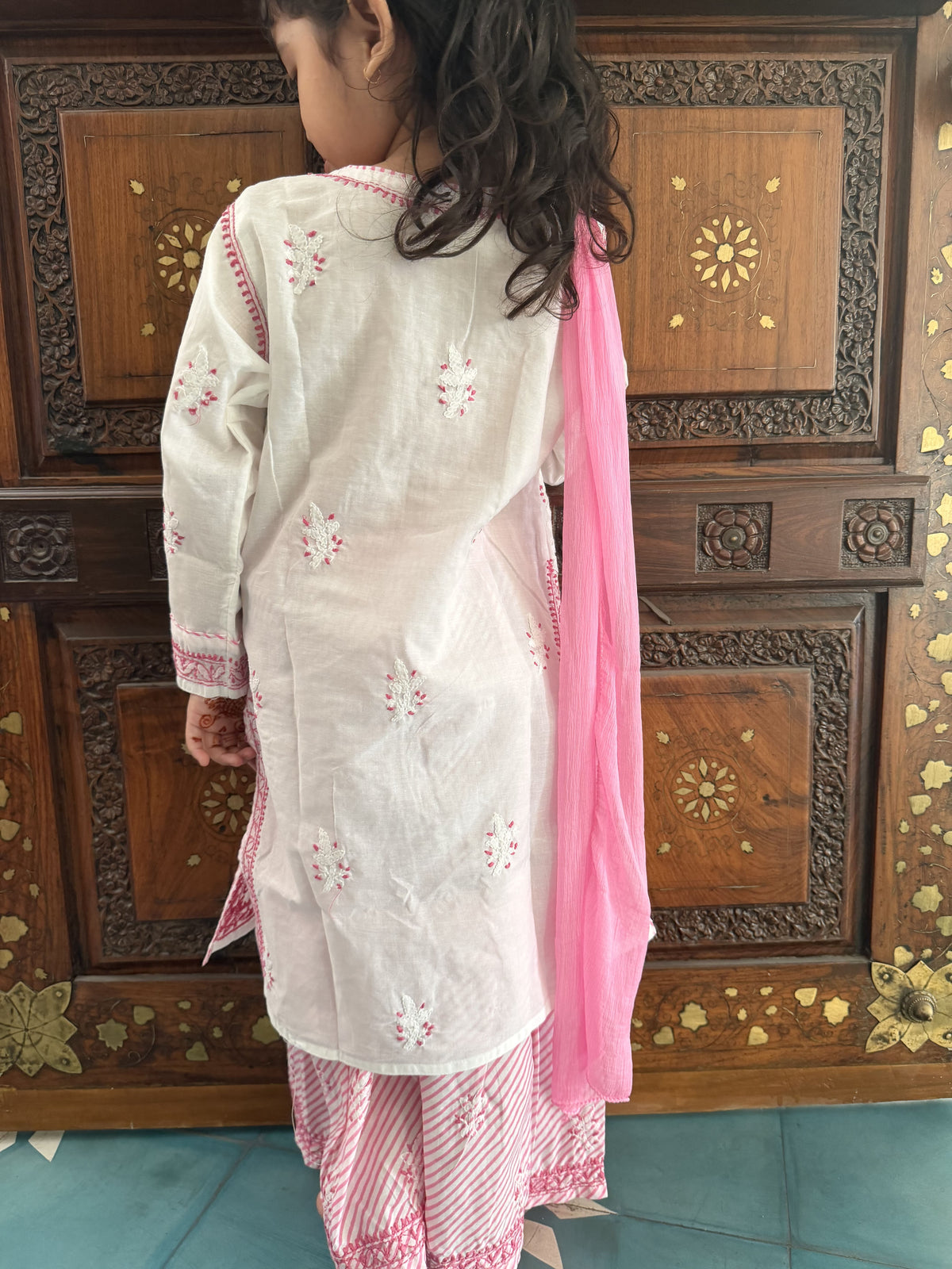 A young girl viewed from behind wearing a white kurta with pink floral embroidery, pink striped pants, and a solid pink dupatta, standing in front of a carved wooden cabinet.
