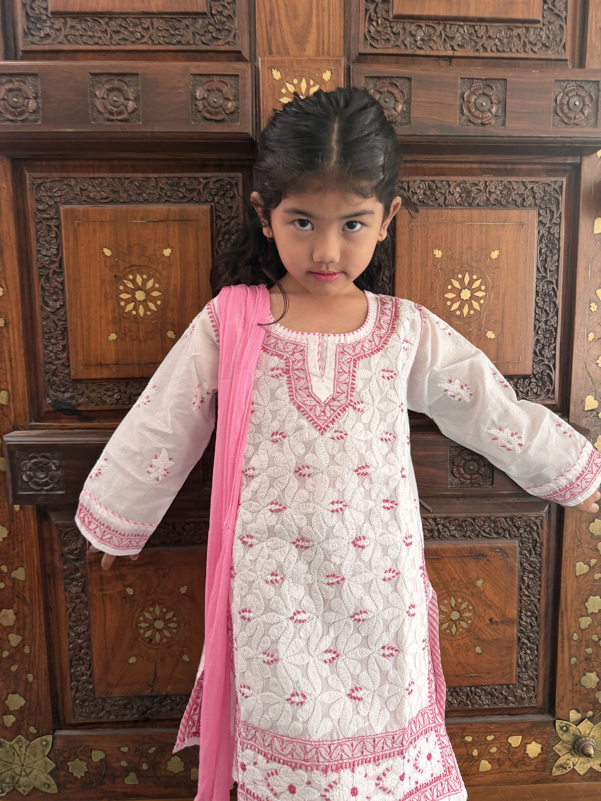A young girl modeling a white embroidered kurta with pink detailing and a pink dupatta, standing in front of a carved dark brown wooden door.