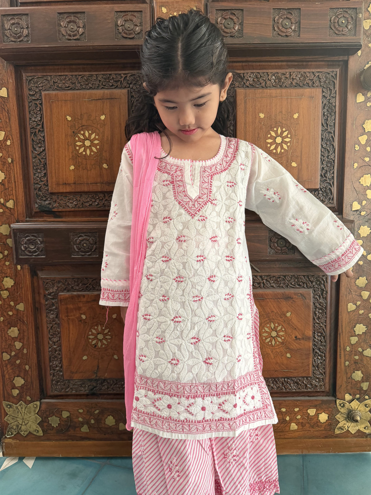 A young girl wearing a traditional white and pink embroidered kurta set with a pink dupatta, standing in front of a carved wooden cabinet.