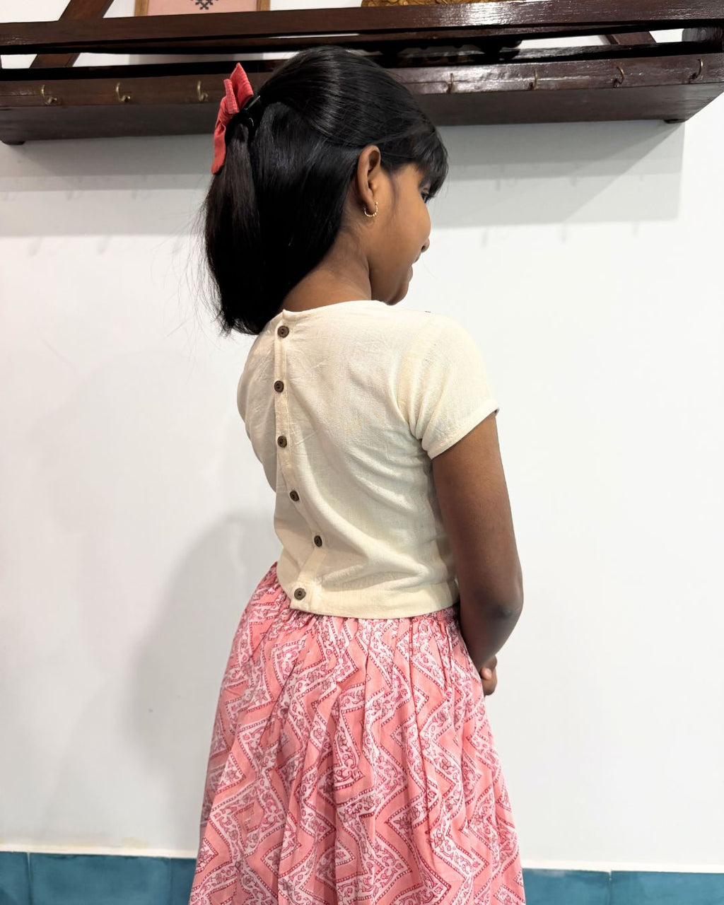 Young girl wearing a white blouse with buttons and a pink patterned skirt, standing against a white wall.