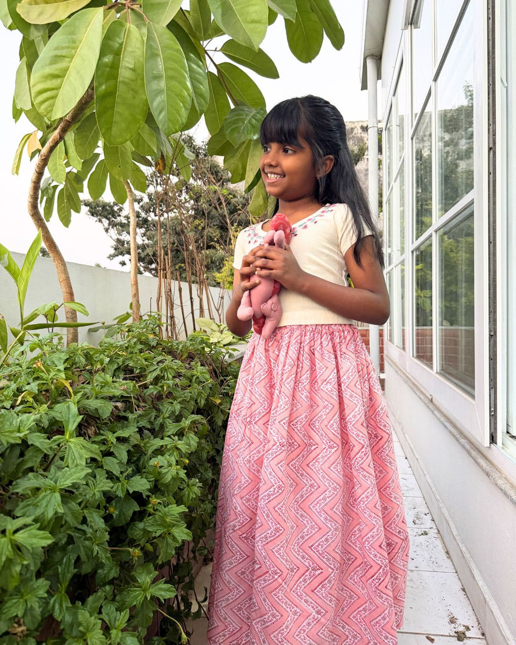 Young girl holding a pink teddy bear outdoors near greenery and a building.