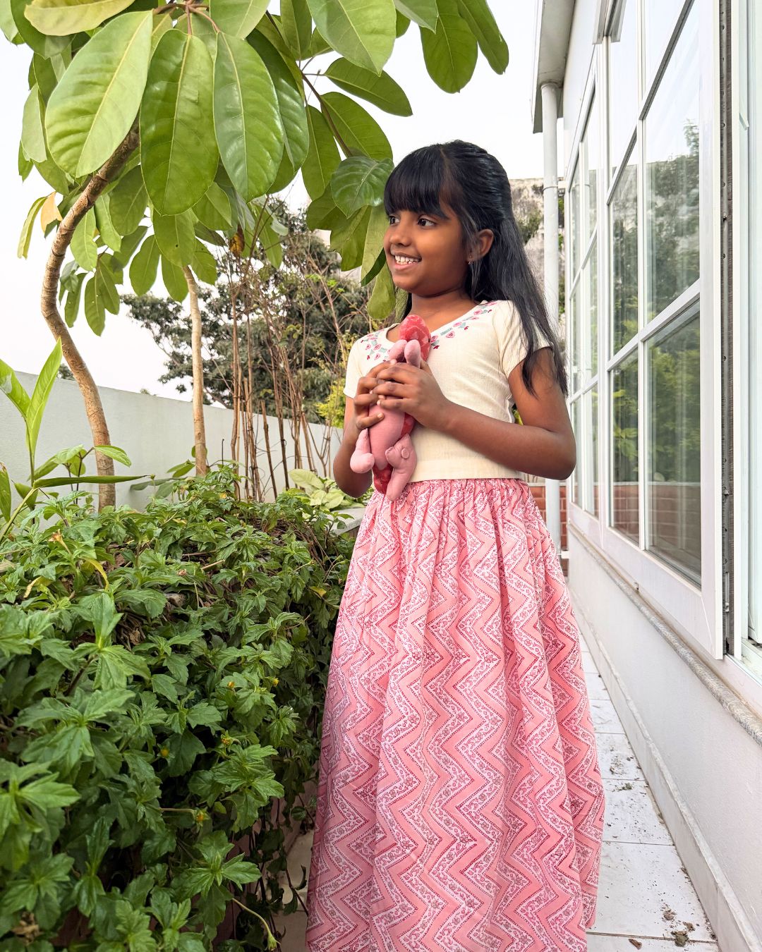 Young girl holding a pink teddy bear outdoors near greenery and a building.