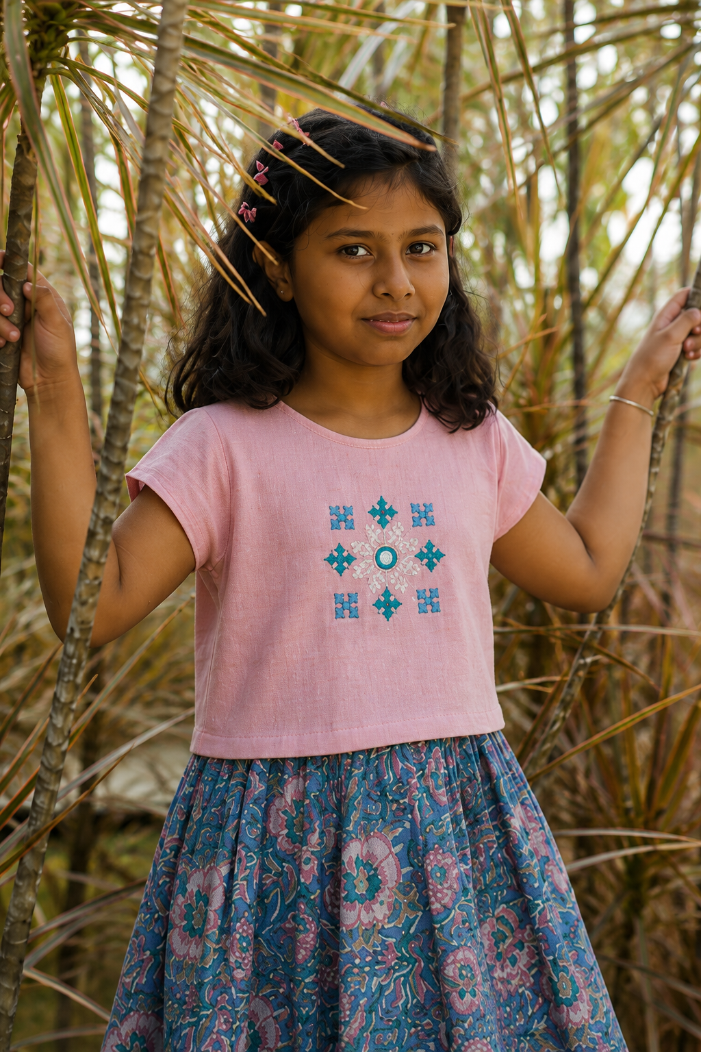 A young girl wearing a light pink short-sleeve top with blue geometric embroidery and a blue floral skirt, standing outdoors among palm trees.
