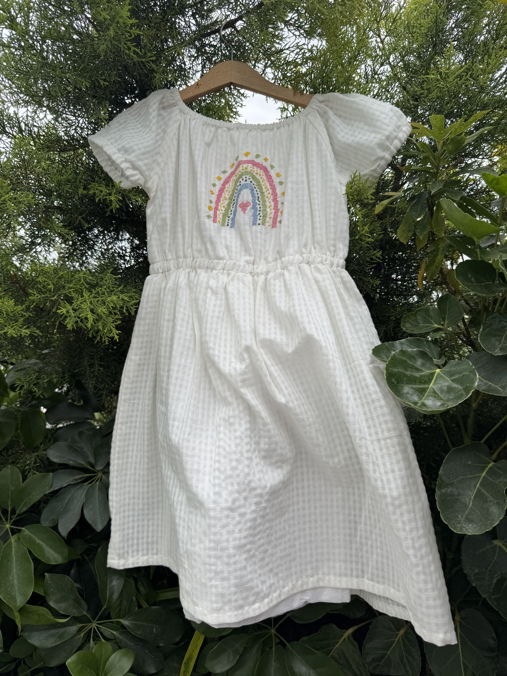 A white textured cotton dress with a colorful rainbow and floral cross stitch embroidery on the chest, hanging outdoors against a background of green leaves.