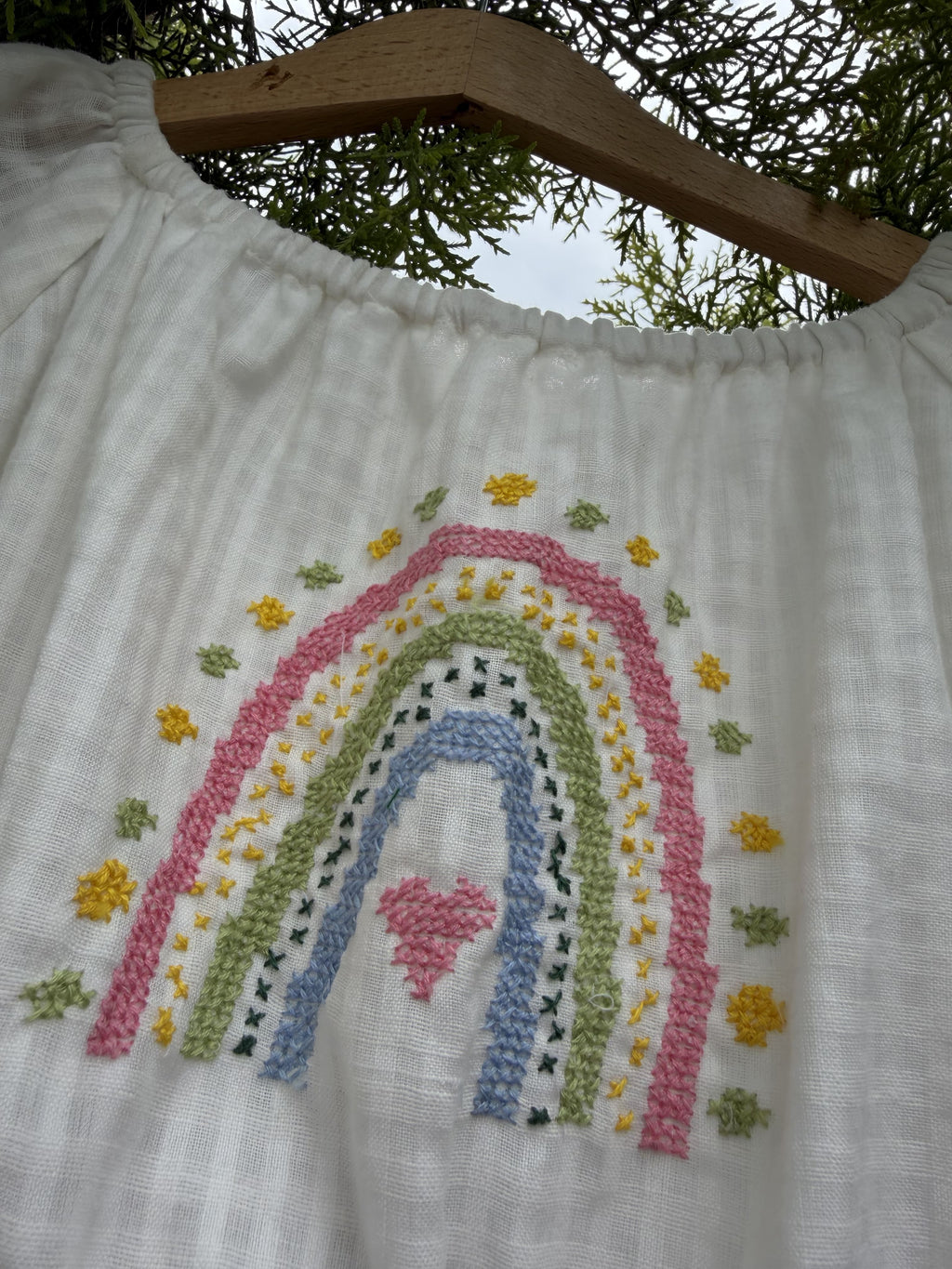 White muslin baby romper featuring a colorful embroidered rainbow and small flowers, shown hanging on a wooden hanger outdoors.