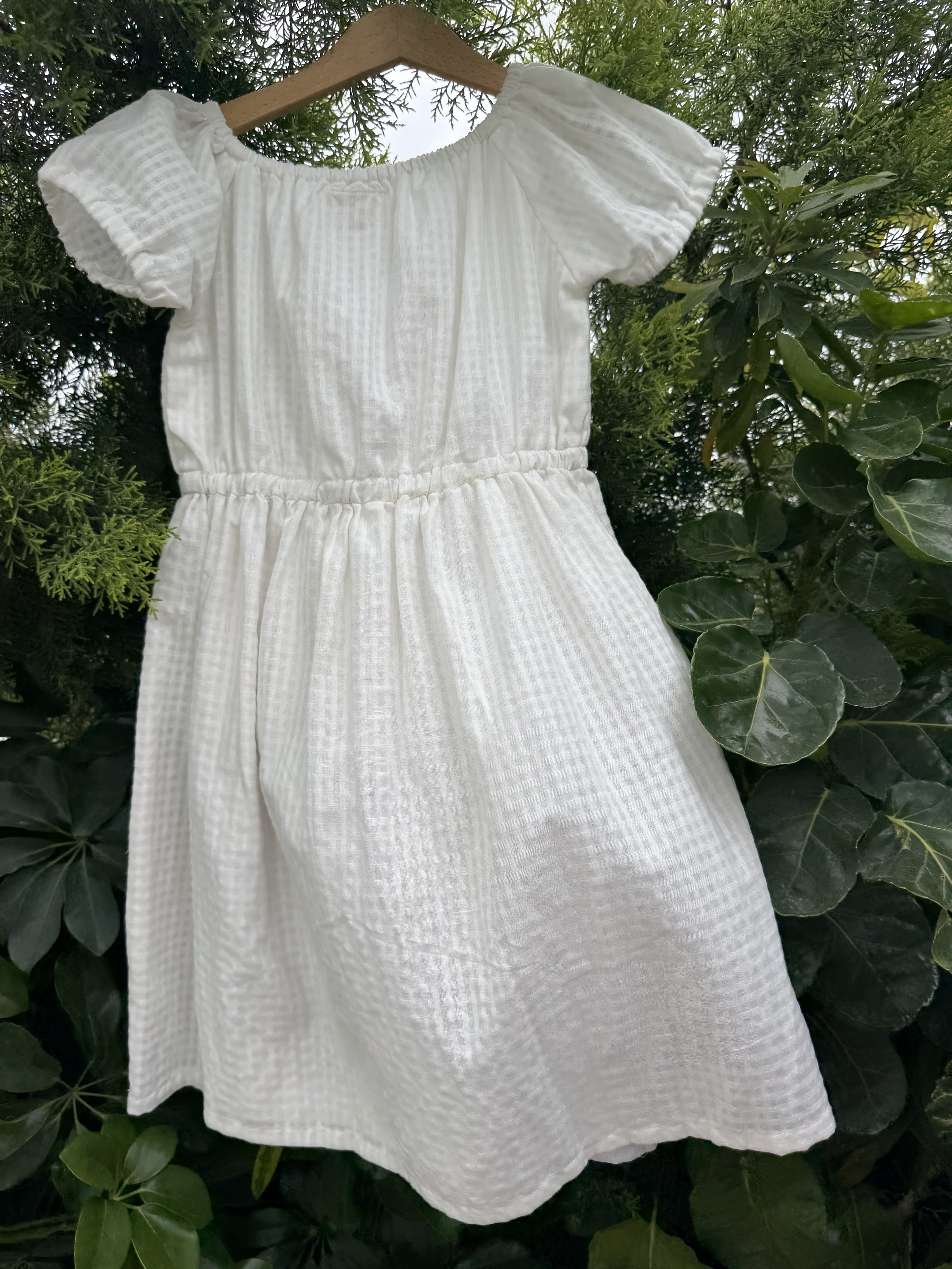 A white textured cotton dress with short puff sleeves and a gathered waist, shown hanging outdoors against a background of green foliage.